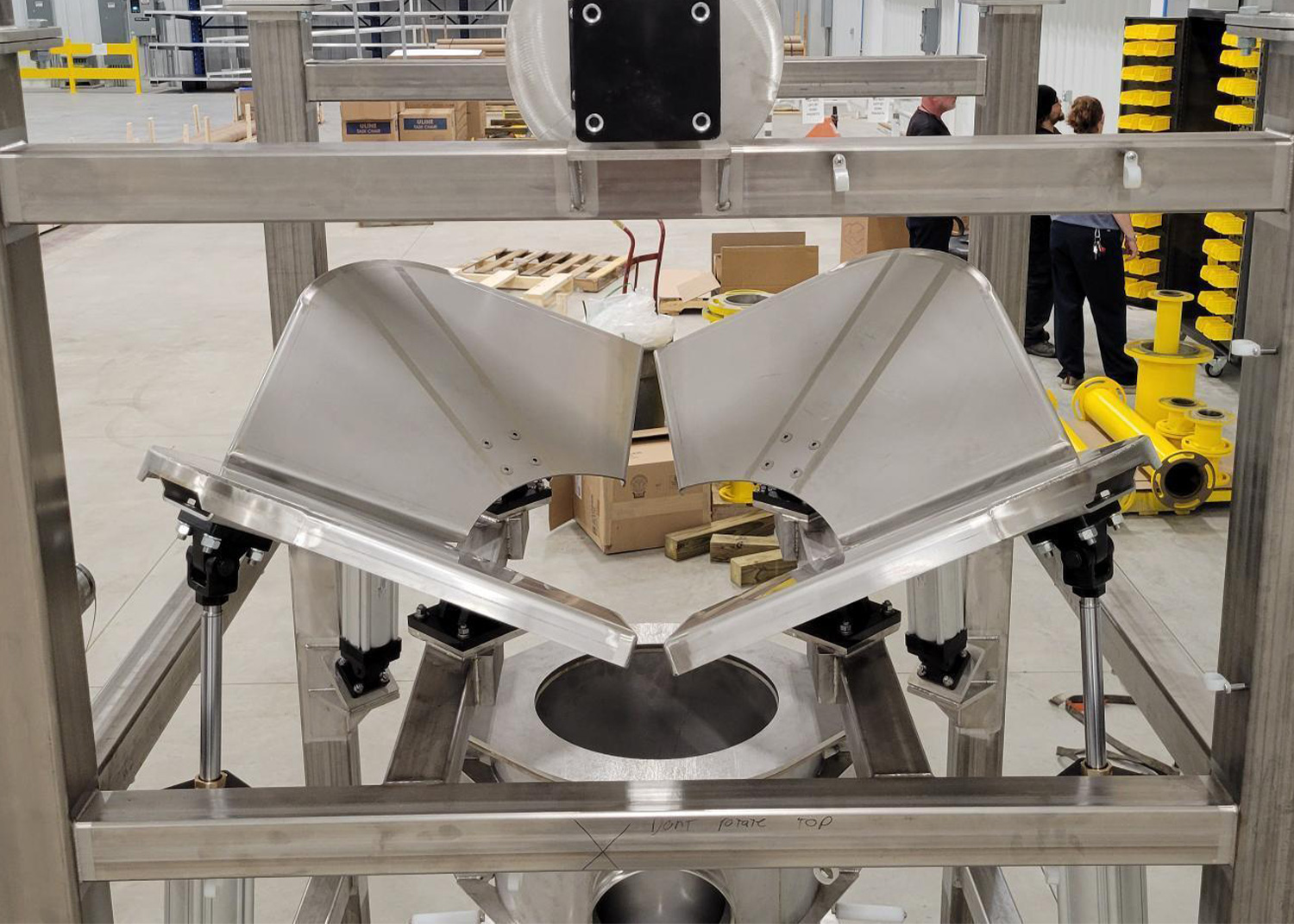 A stainless steel industrial machine featuring two adjustable V-shaped metal gates positioned above a circular opening in a warehouse setting.