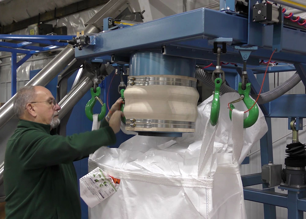 A worker attaches the loops of a large white bulk bag to the hooks of an industrial filling machine.